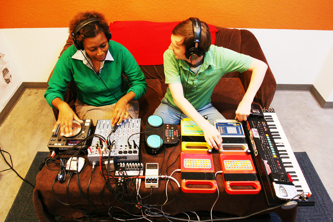 Beatrix Jar portrait, both sitting on brown sofa with red blanket hanging over back. Both are wearing green shirts and headphones. A table sits before them with electronic instruments displayed. Both are playing. 